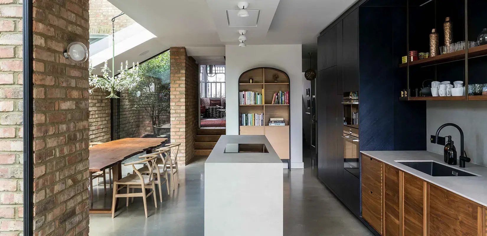 Open-plan kitchen and dining extension with exposed brick piers, a polished concrete floor, a white island, dark blue and walnut cabinetry, an arched oak bookcase and a glazed side return opening onto a planted courtyard.