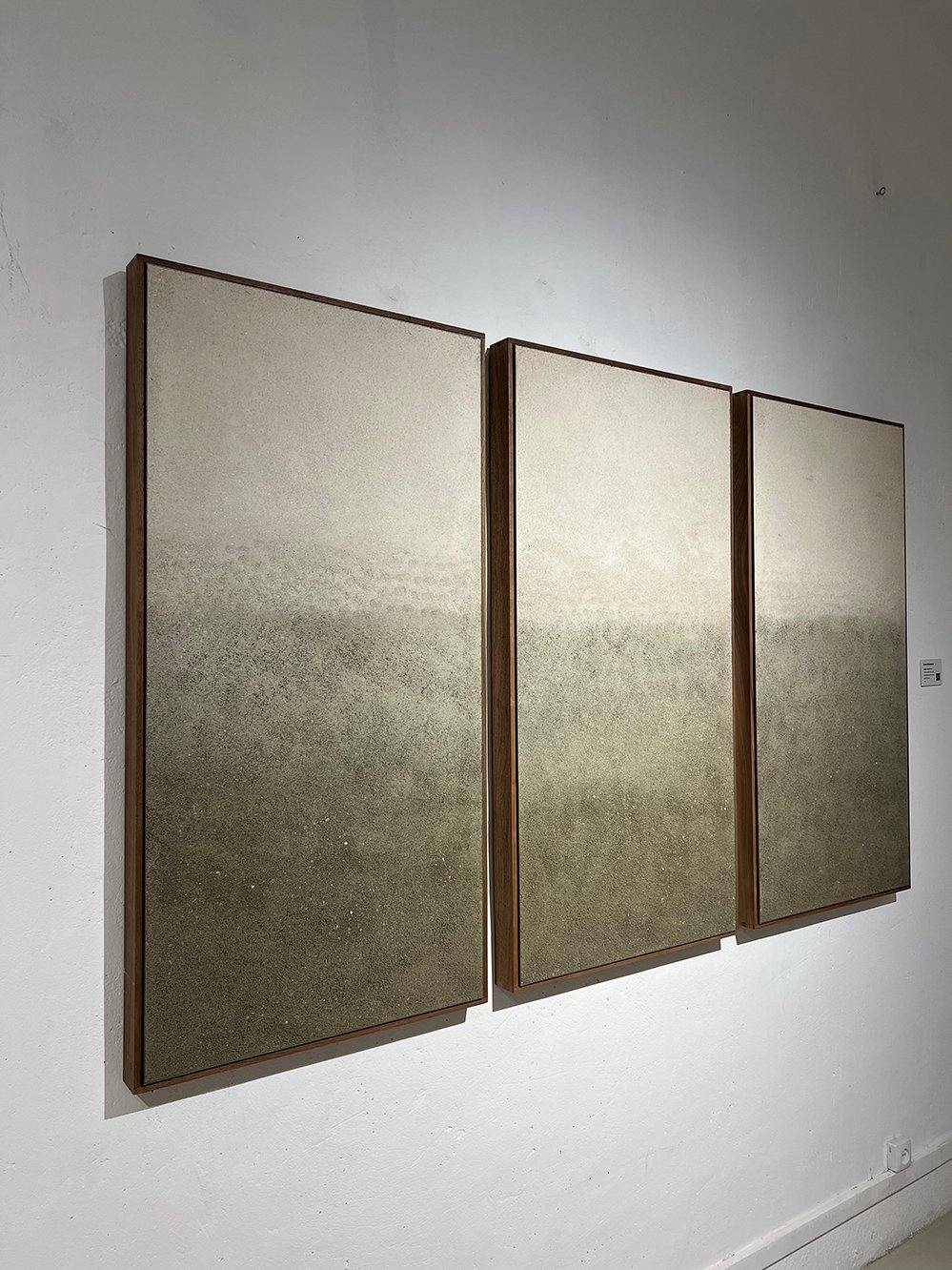 Three framed clay and mineral pigment panels by Adam Weismann arranged side by side, showing a graduated tonal shift from pale cream at the top to deep olive grey at the base. Claymoon Studio, Matiz Gallery London. 2026.
