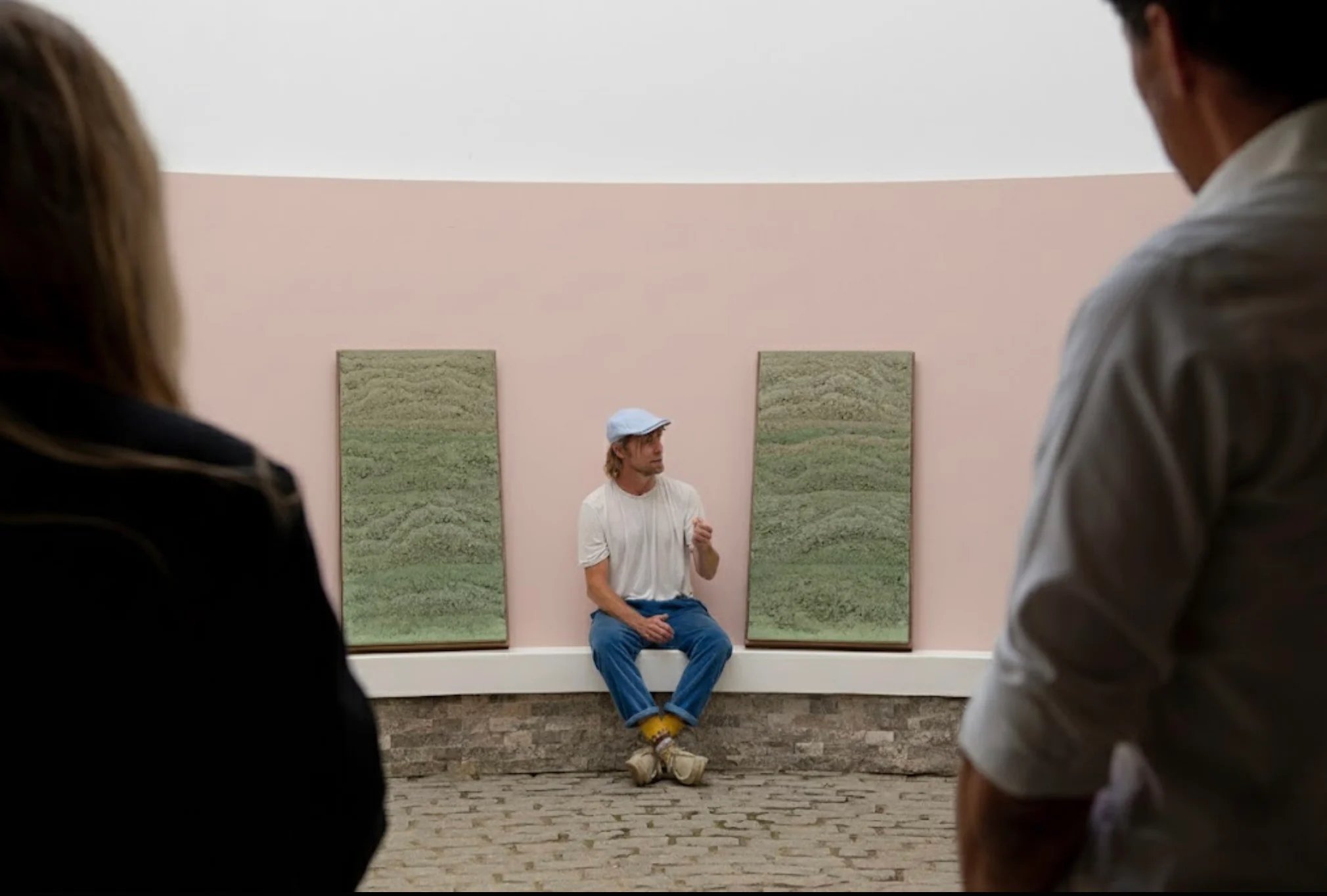Adam Weismann of Clayworks sits on a low ledge against a curved pink wall, between two tall textured green clay artworks resembling layered sediment, speaking to two people seen from behind in the foreground of a gallery space with a cobbled floor. RISE Design Studio.