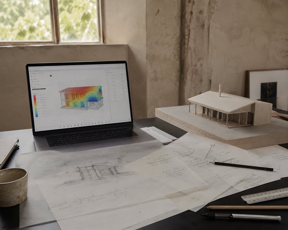 Architect's studio desk with laptop showing building energy model heat map, hand-drawn pencil sketches on trace paper, timber scale model and drawing tools in natural light.