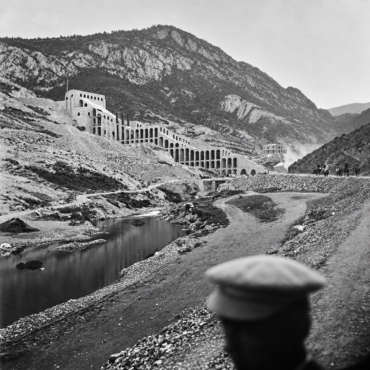 Black and white archive photograph of the Asland cement factory at the Clot del Moro under construction, tiers of Catalan brick vaults stepping down a bare limestone hillside above the Llobregat, with chimneys, riders on the track and a worker in a flat cap in the foreground. RISE Design Studio.