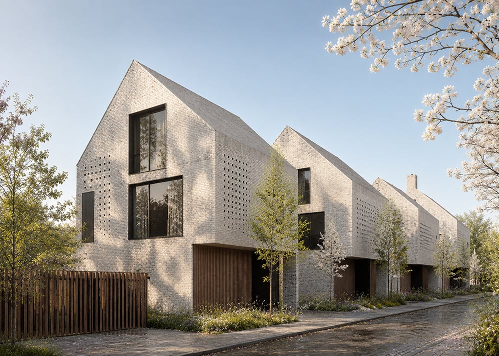 A terrace of contemporary gable-fronted houses in pale grey brick, with perforated brickwork panels, full-height windows and recessed timber-clad entrance bays, set behind young trees and planting along a cobbled street in blossom. RISE Design Studio.