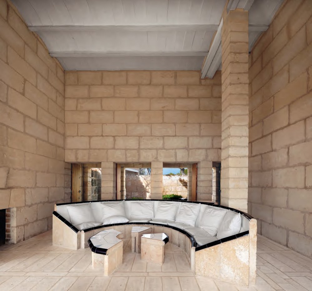 Living room at Can Lis by Jørn Utzon with a curved built-in stone bench edged in black tiles and white cushions, hexagonal stone tables, tall marés walls and small openings to the courtyard. RISE Design Studio.