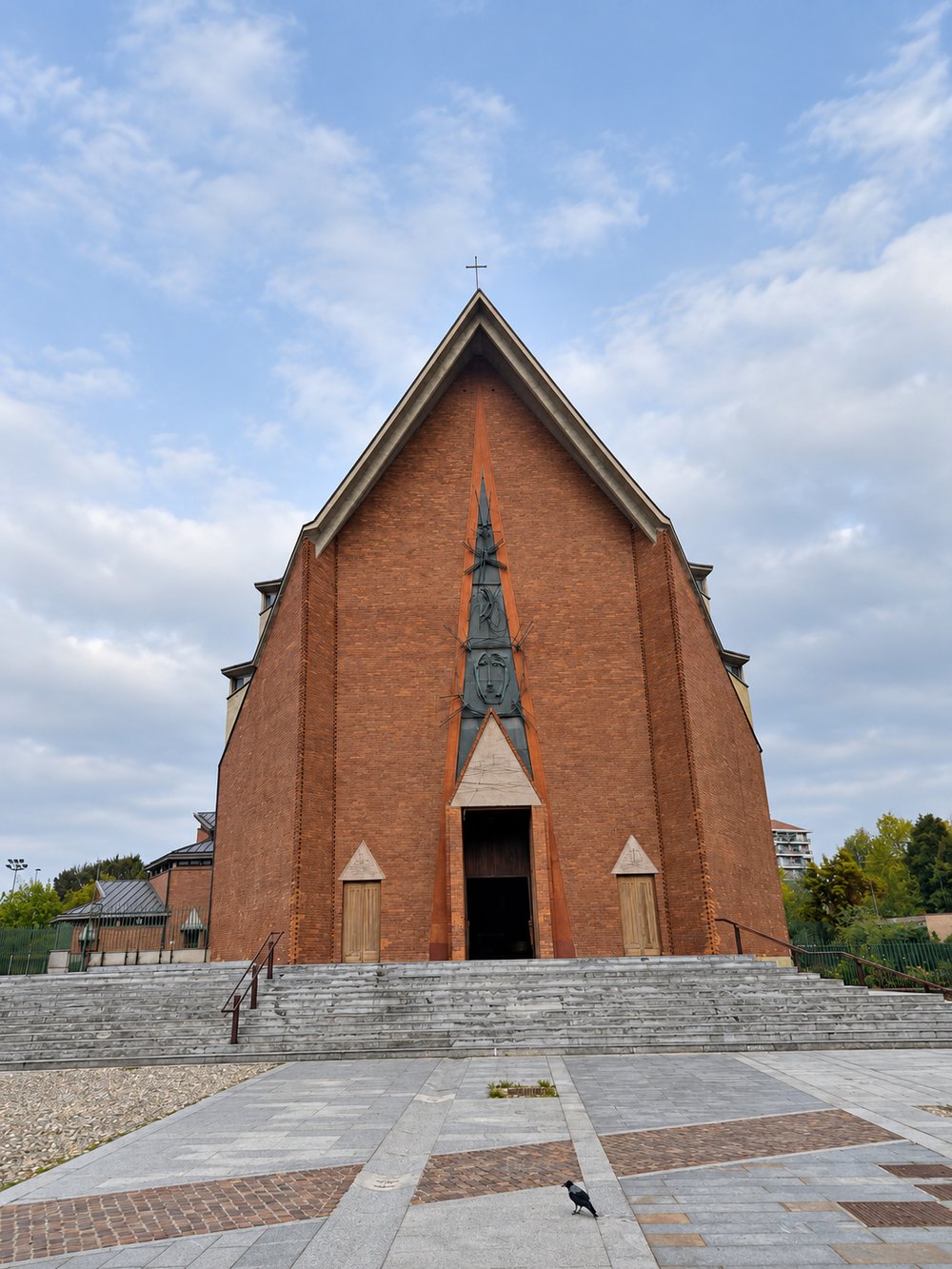 Entrance front of the Chiesa del Gesù Redentore, Turin: a tall red brick gable with a narrow lancet holding a sculpted metal panel, a triangular stone tympanum over the timber doors, two side doors, and wide stone steps, with a crow on the paving. RISE Design Studio.