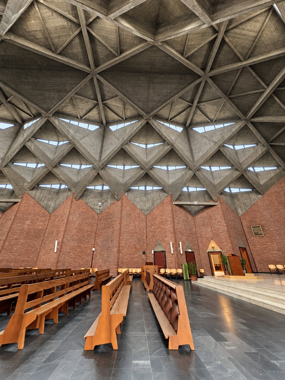 Raking view up a folded red brick wall inside the Chiesa del Gesù Redentore, Turin, with a cross picked out in projecting bricks, meeting a board-marked concrete vault with a triangular rooflight slot. RISE Design Studio.