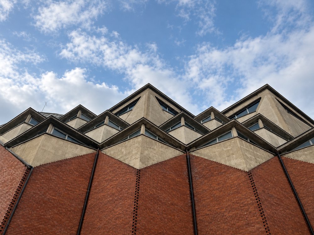 Interior of the Chiesa del Gesù Redentore, Turin, looking up at a board-marked concrete vault of triangular ribs, with rows of glazed triangular pockets along its base above zigzag red brick walls, timber pews and steps to the altar. RISE Design Studio.