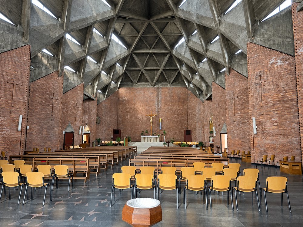 Wide view of the nave of the Chiesa del Gesù Redentore, Turin, looking to the altar: a faceted concrete vault of triangular ribs with rooflights, folded red brick walls with brick crosses, timber pews, yellow chairs, a font and a black stone floor with pink triangle inlays. RISE Design Studio.