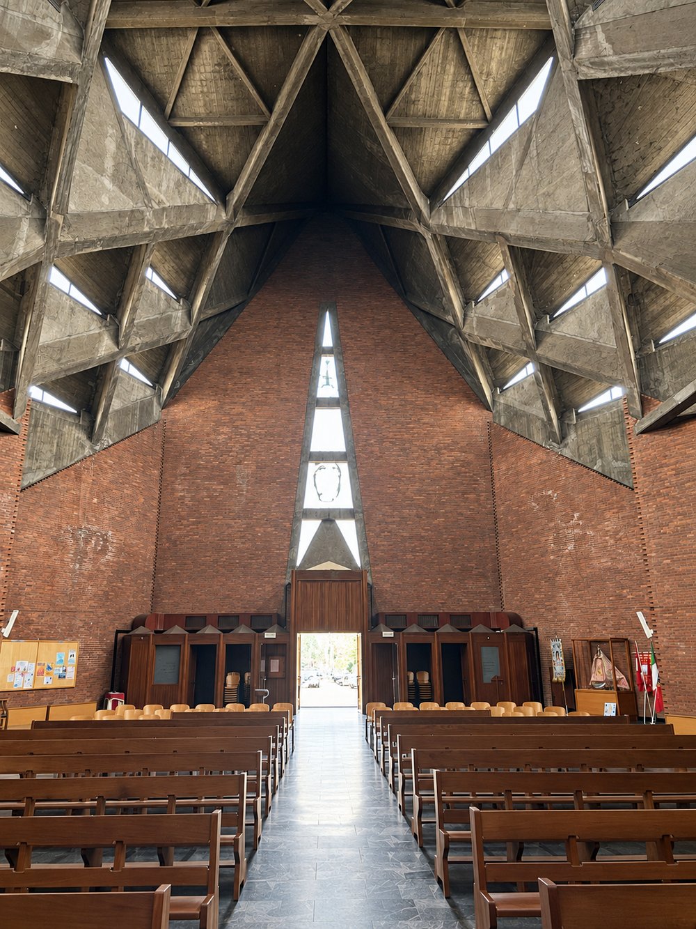 Nave of the Chiesa del Gesù Redentore, Turin, looking back to the entrance: a tall red brick gable wall with a narrow lancet window of stacked triangular panes above timber doors, pews either side and a faceted concrete vault with rooflights overhead. RISE Design Studio.
