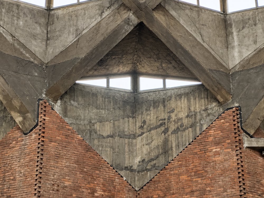 Close view inside the Chiesa del Gesù Redentore, Turin, of a board-marked concrete rooflight pocket with a horizontal slot of daylight, its triangular facet landing on the V-shaped fold of a red brick wall below. RISE Design Studio.