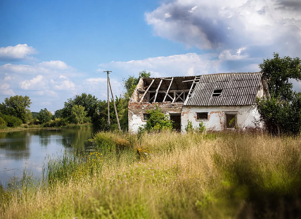 Derelict single-storey farmhouse in rural Kent with crumbling brick walls, a collapsing roof and overgrown vegetation, photographed before its replacement by a new rammed earth dwelling designed by RISE Design Studio.