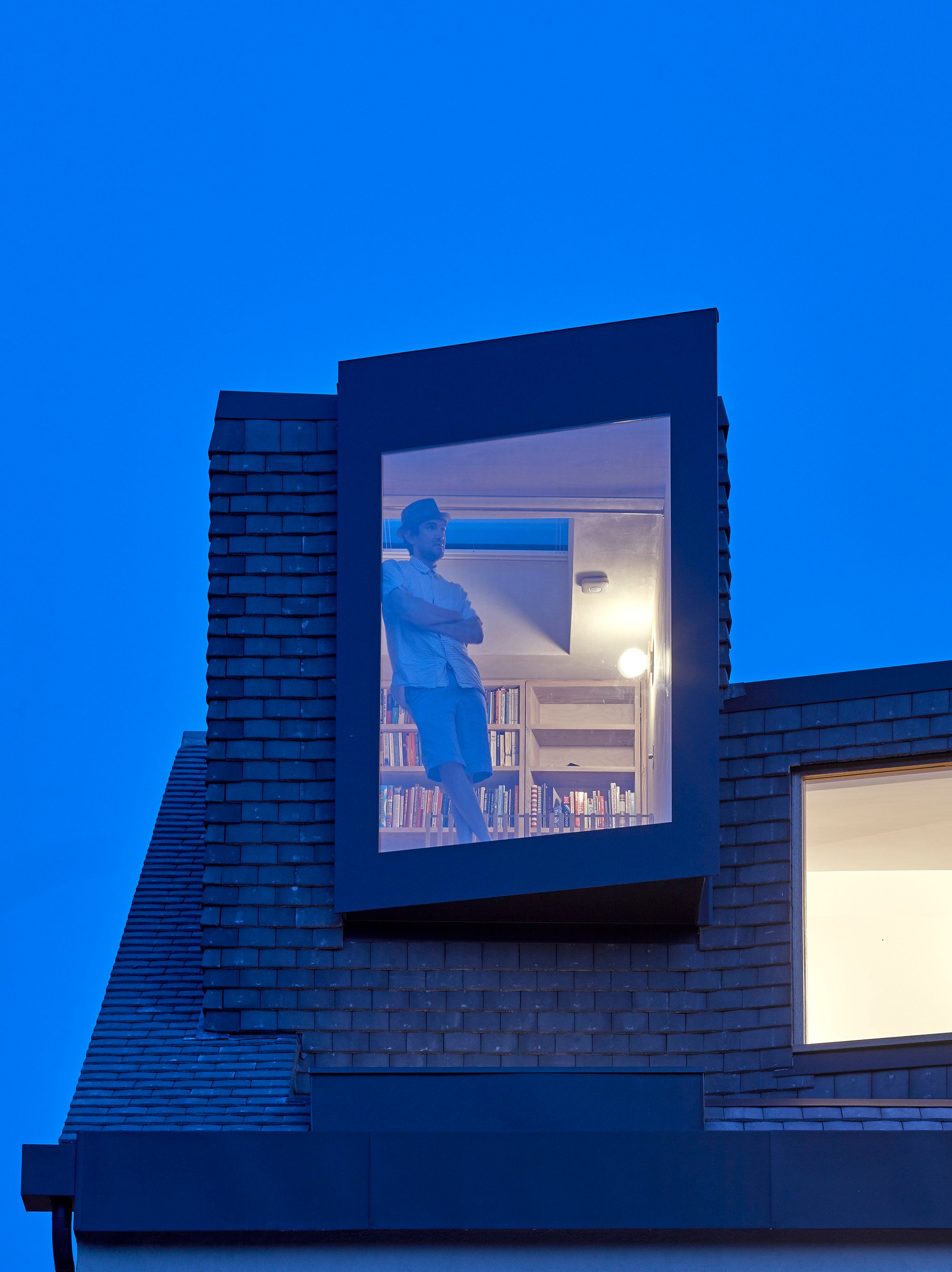 Dusk view of a slate-clad dormer at Douglas House by RISE Design Studio, with a projecting dark metal oriel window and an opening rooflight in the ceiling behind it for stack ventilation. A man leans against a bookshelf inside the lit loft study, deep blue evening sky above. RISE Design Studio.