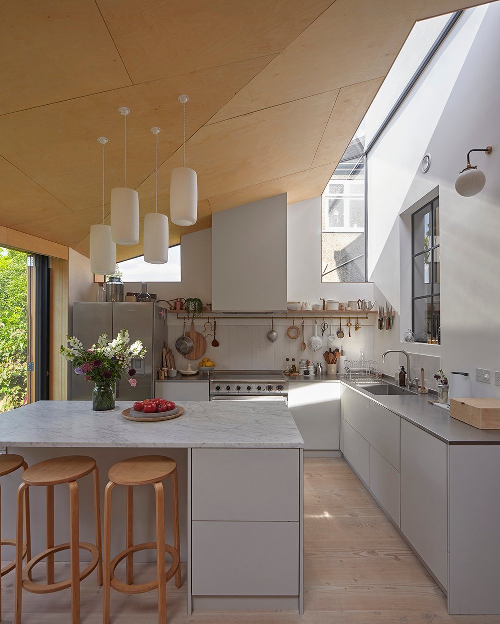 Douglas fir-lined kitchen at Douglas House in Kensal Rise by RISE Design Studio, with white pendant lights, a marble island, a range cooker and clerestory skylights drawing daylight across the timber ceiling toward sliding doors that open onto the garden.