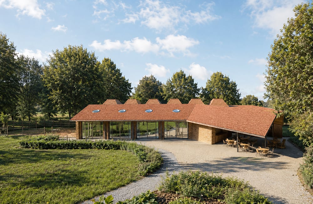 Contemporary low-energy tennis club pavilion in Kensal Rise, London, featuring red tiled pitched roofs, circular rooflights, exposed brickwork, large glazed openings, covered terraces, and landscaped gardens designed by RISE Design Studio