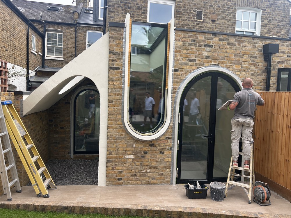 Rear of Flower House, a Victorian terrace in Queen's Park, during final works: a sweeping white lime-rendered canopy over a lightwell, a tall curved window and arched garden doors set in London stock brick. RISE Design Studio.