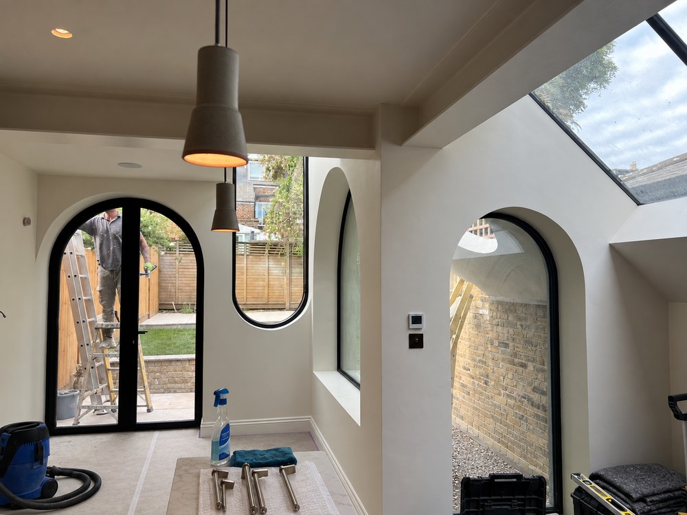Kitchen extension at Flower House nearing completion, with arched black-framed garden doors, a curved window, an arched lightwell window and roof glazing, and the pendant lights switched on. RISE Design Studio.