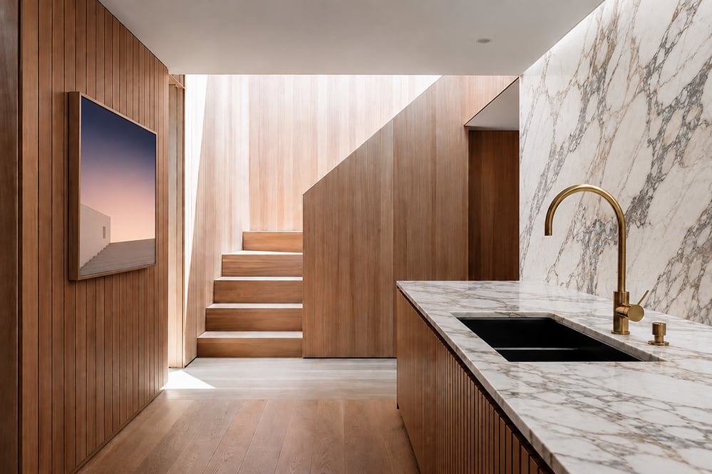 Interior of a renovated London mews house showing a timber-clad staircase lit by a rooflight shaft, marble kitchen worktop with brass tap, and slatted oak panelling throughout.