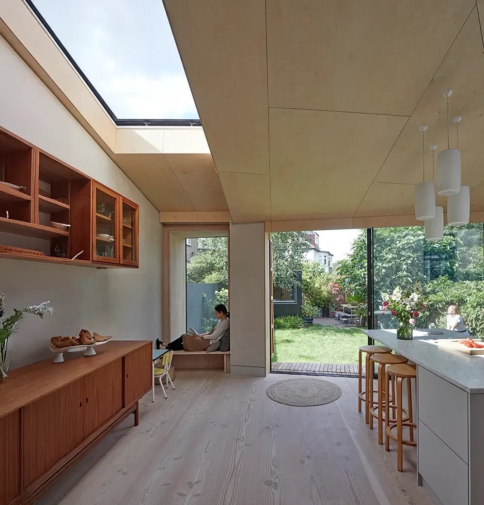 A calm, light-filled retrofit kitchen with a plywood-lined ceiling, a long rooflight and a built-in window seat where a person reads. Pale timber floor, mid-century teak sideboard and a marble-topped island, with full-height glazing opening to a green garden beyond. RISE Design Studio.