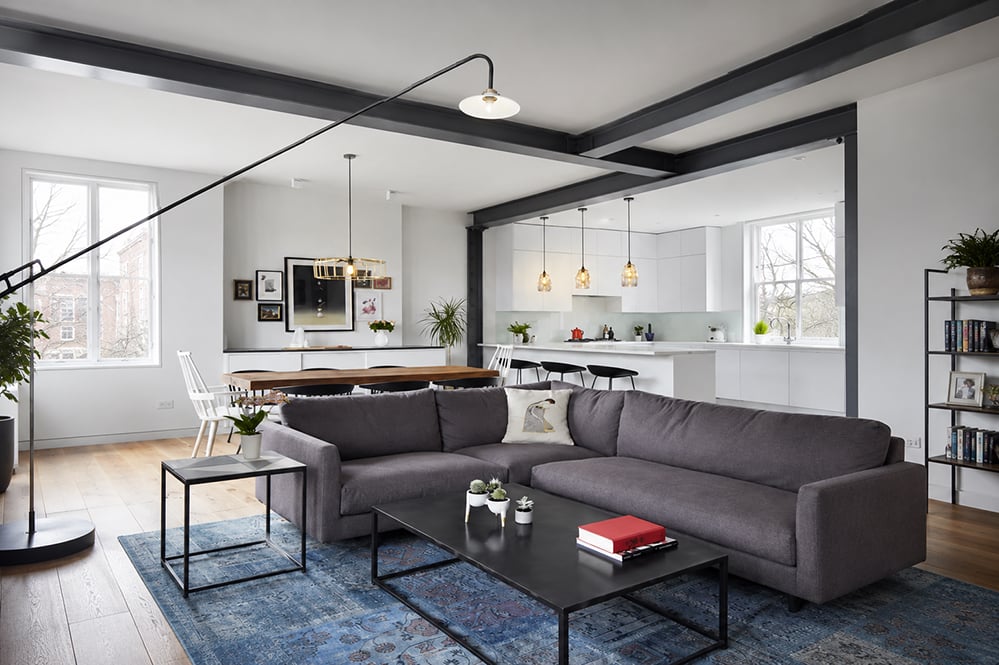 Open-plan living and kitchen in a renovated North London Victorian home, featuring exposed steel beams, oak flooring, grey corner sofa, pendant lighting and views to mature garden trees beyond.