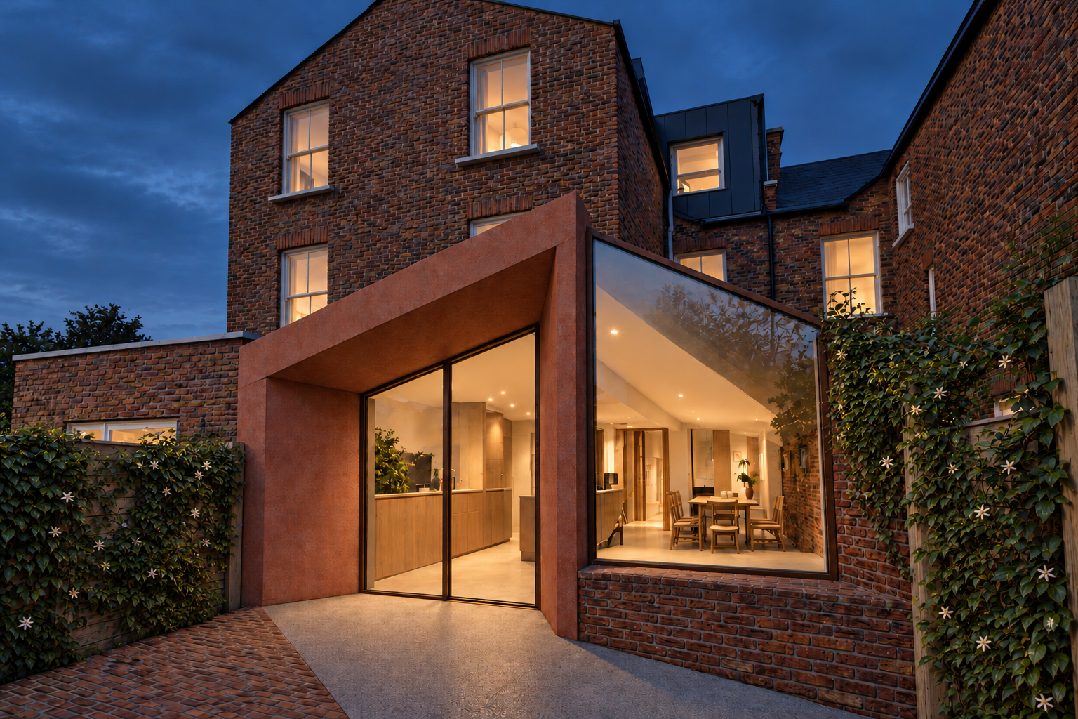 View at dusk of a folded, terracotta-toned rear extension to a Victorian brick terrace, with a glazed corner looking into the kitchen and dining space