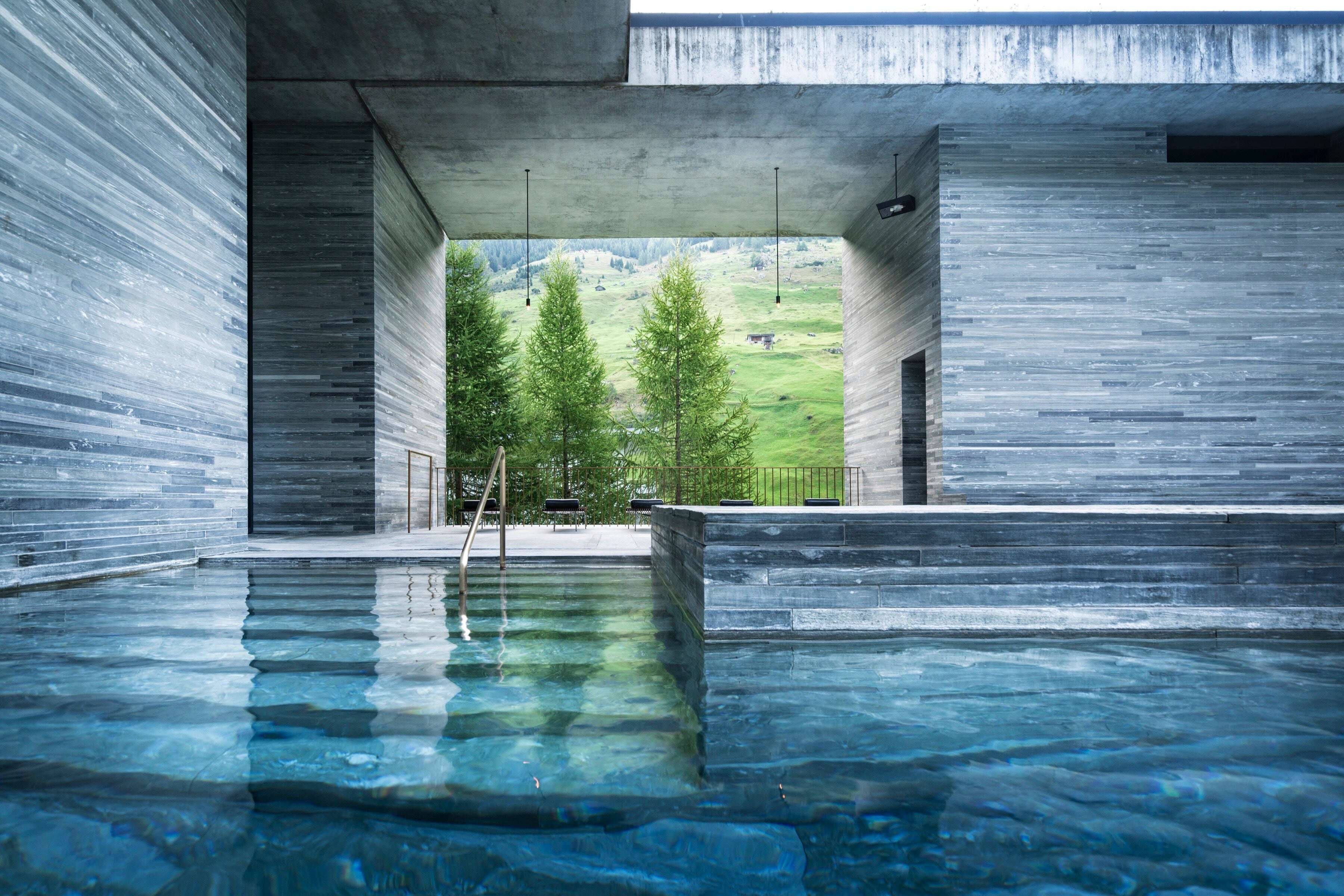 Indoor thermal pool at Peter Zumthor's Therme Vals in Switzerland, with layered grey quartzite walls, a board-marked concrete ceiling and a brass handrail leading into pale blue water, and a wide opening framing larch trees and a green Alpine hillside beyond. RISE Design Studio.