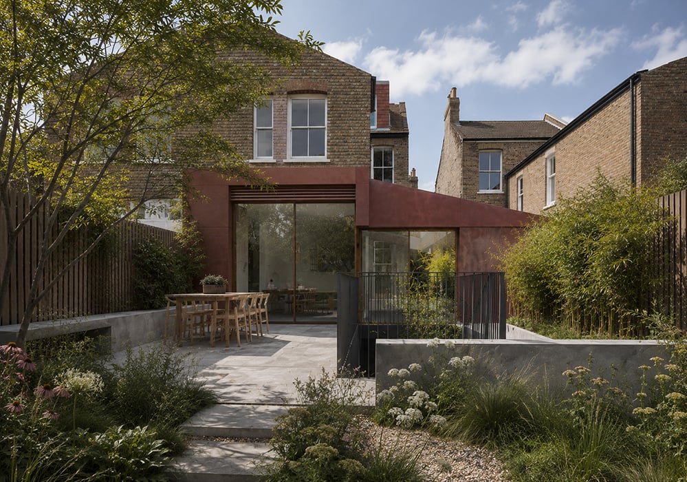 A rear extension to a Victorian brick house, clad in deep red-brown render with a projecting roof canopy and full-height glazing shading the interior, opening onto a stone terrace with a dining table and a densely planted garden. RISE Design Studio.