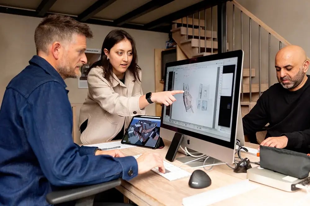 Three architects at RISE Design Studio reviewing a BIMx 3D building model on an iMac screen and iPad tablet during a collaborative design meeting in their London studio