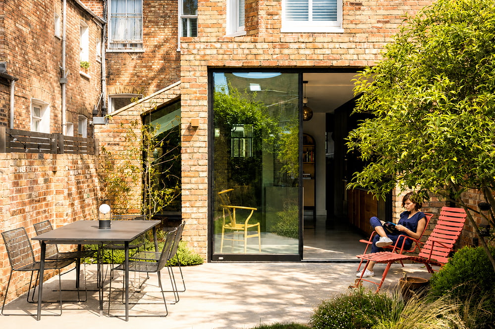 Rear garden view of a contemporary brick house extension in Queen’s Park, London, designed by RISE Design Studio with large sliding glass doors, reclaimed brickwork, polished concrete terrace, and landscaped planting.
