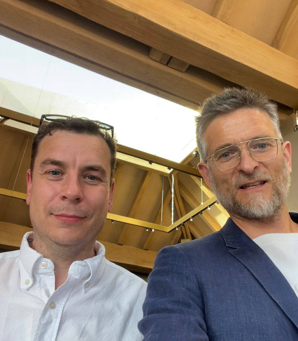 Two men beneath the oak roof structure and glazed rooflight of the Department Store's rooftop level in Brixton, one in a white shirt, one in a navy linen blazer, with the timber frame rising behind them. RISE Design Studio.