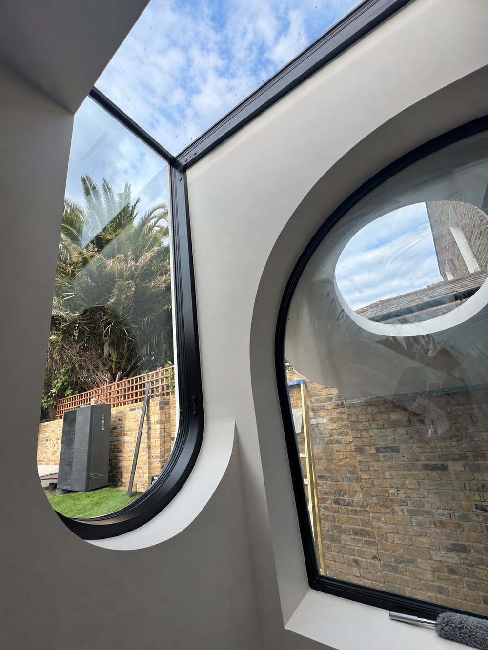 Inside the Flower House extension, an arched window with a deep curved reveal looks onto the lightwell and the canopy's circular opening, beside a curved window rising into roof glazing towards garden planting. RISE Design Studio.