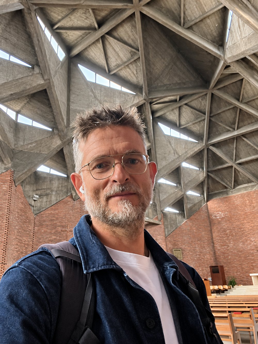 Sean Ronnie Hill of RISE Design Studio, in glasses and a dark denim jacket, photographed from below inside the Chiesa del Gesù Redentore, Turin, with the triangular concrete vault, rooflights and red brick walls behind him. RISE Design Studio.