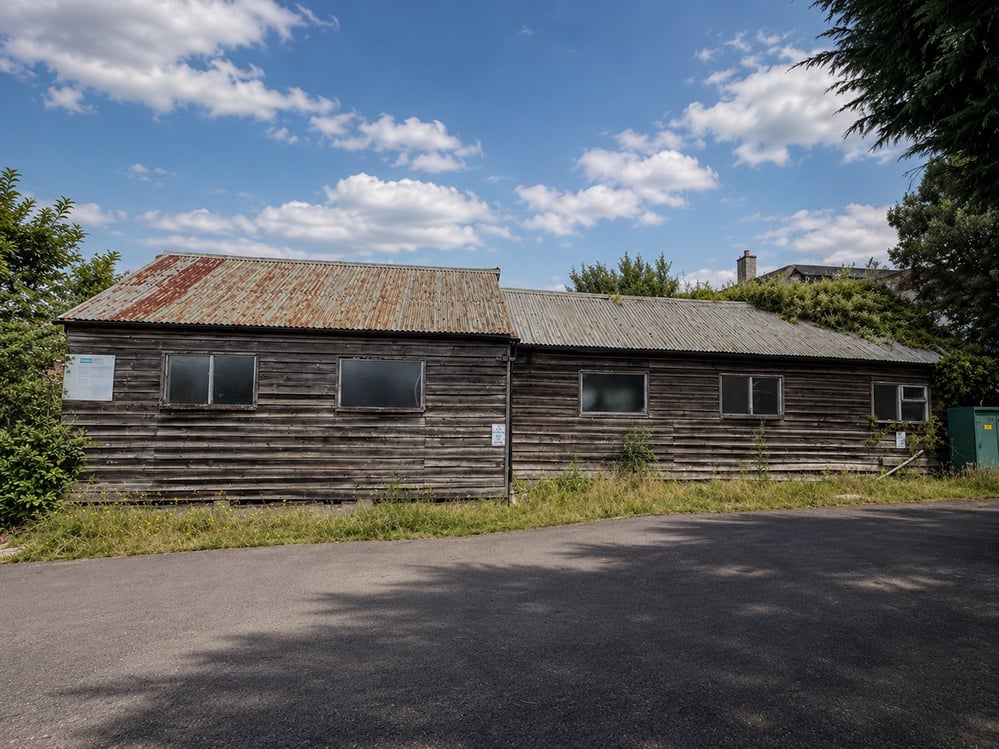 A long, single-storey weathered timber-clad tennis pavilion with a rusting corrugated-metal roof, small windows and overgrown planting climbing one end, set behind a tarmac forecourt under a blue sky. RISE Design Studio.