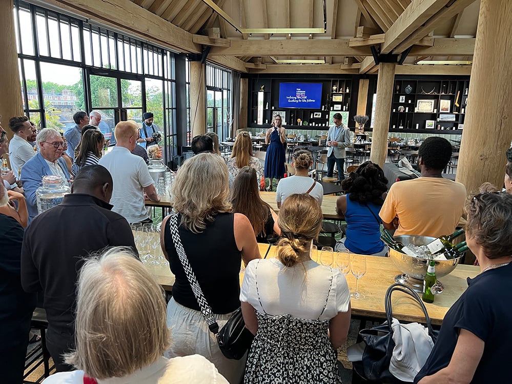 Guests at a London Society drinks reception fill the green-oak-framed rooftop room of the Department Store in Brixton, facing a speaker holding a microphone. Tall Crittall windows look over Brixton rooftops, with a dark bar and curated shelving behind and drinks on long oak tables. RISE Design Studio.