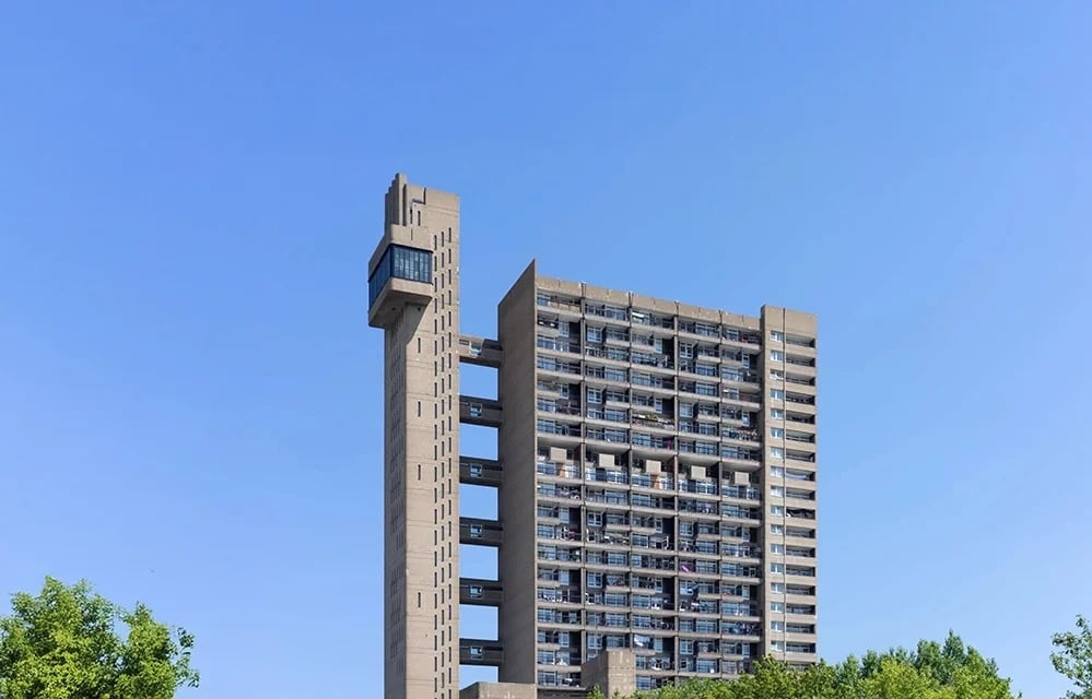 Trellick Tower in North Kensington against a clear blue sky: a tall Brutalist concrete residential block with a separate slender service tower linked by walkways, balconies along the main façade, and green treetops at the base. RISE Design Studio.