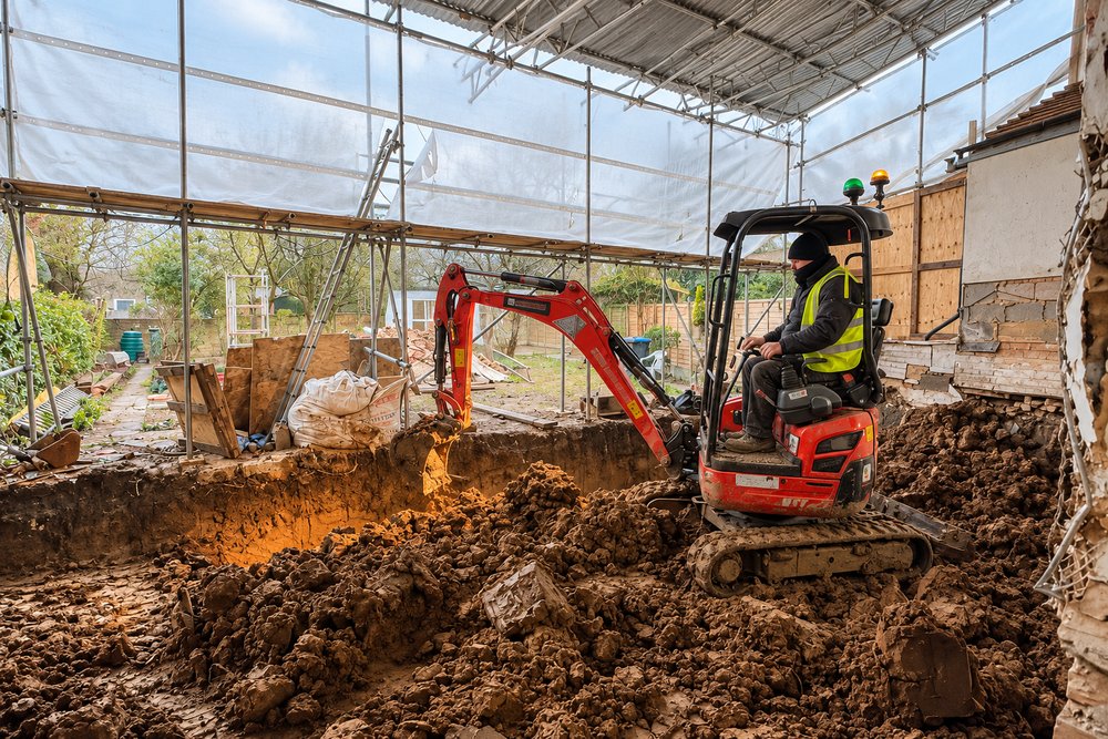 Mini excavator digging foundations for a rear extension under a scaffold canopy, with the existing brick wall of a Victorian house exposed on the right.