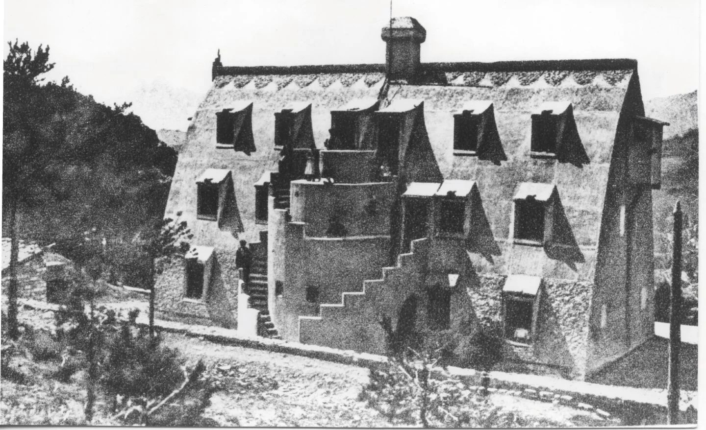 Black and white archive photograph of the newly completed Xalet del Catllaràs around 1905, showing the pebble-dashed catenary roof-walls, projecting dormer hoods, corbelled chimney, the semicircular external stair with people sitting on it and a horse at the foot. RISE Design Studio