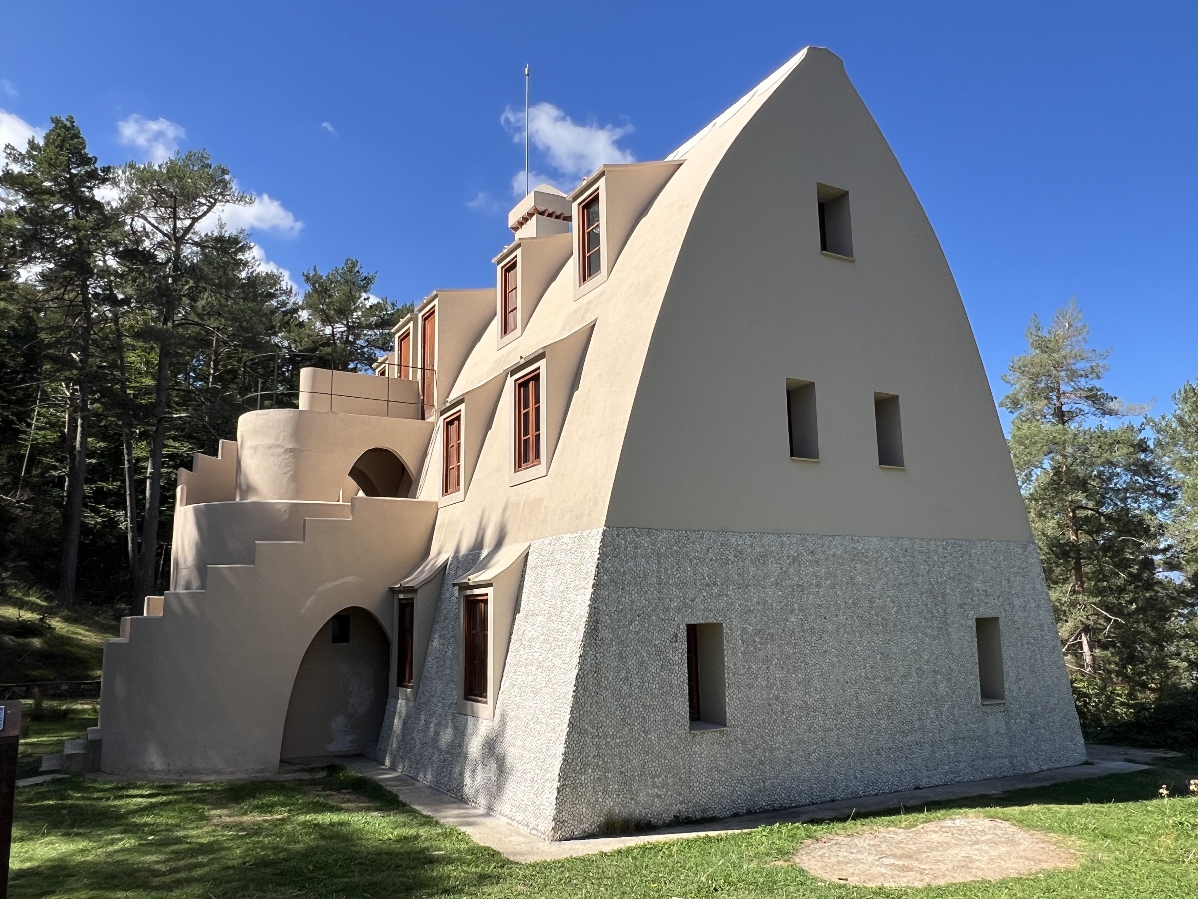 Three-quarter view of the Xalet del Catllaràs, a tall pointed catenary-section house in pale lime render with a white river-pebble base, dormer windows stepping up the roof, small openings in the gable end and a rendered external stair, set in a pine-fringed meadow under a blue sky. RISE Design Studio