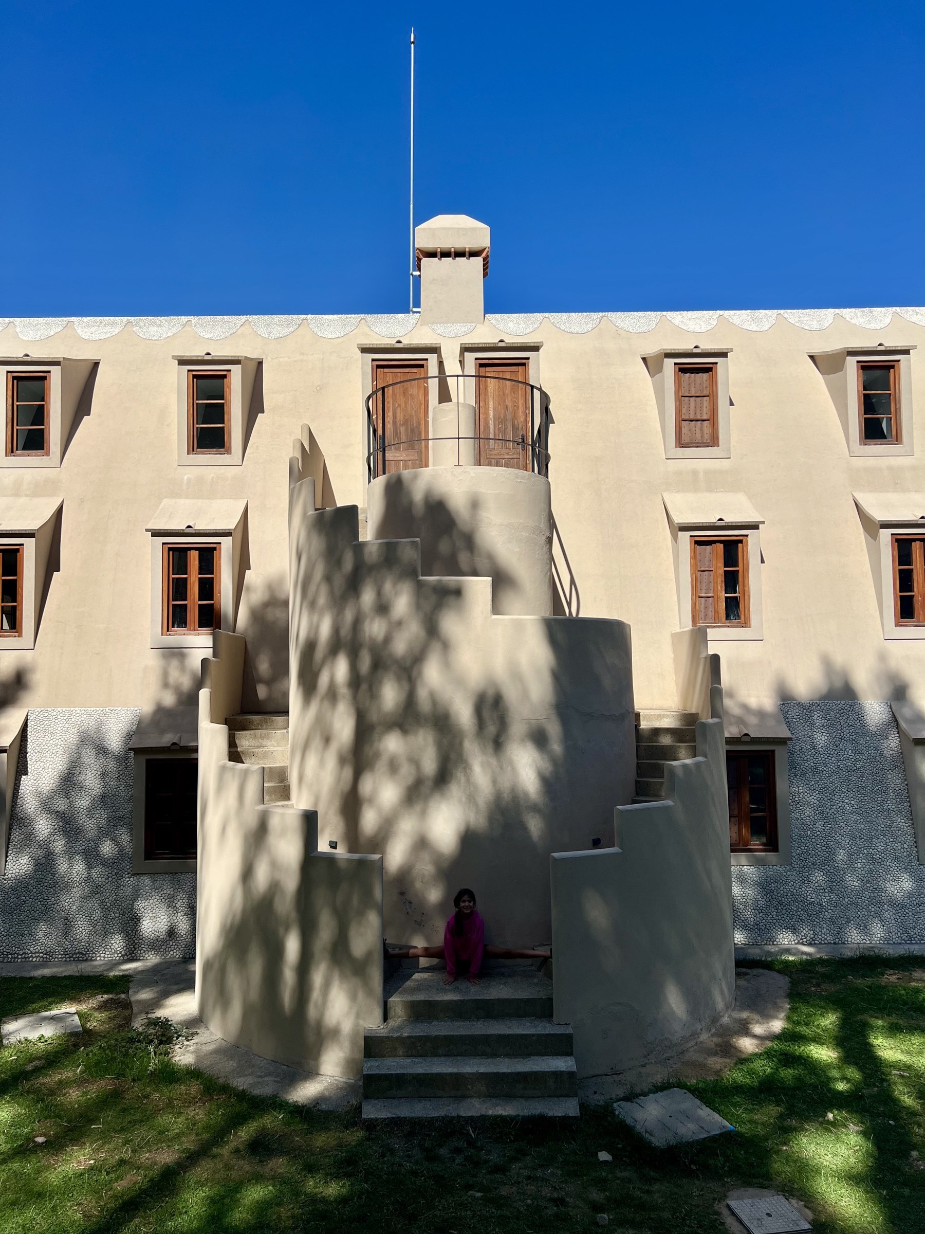 Front elevation of the Xalet del Catllaràs showing the rebuilt semicircular rendered staircase rising in stepped drums to a curved landing with an iron rail, flanked by rows of dormer windows with timber frames and a pebble-dashed ground floor, with dappled tree shadow across the render. RISE Design Studio.