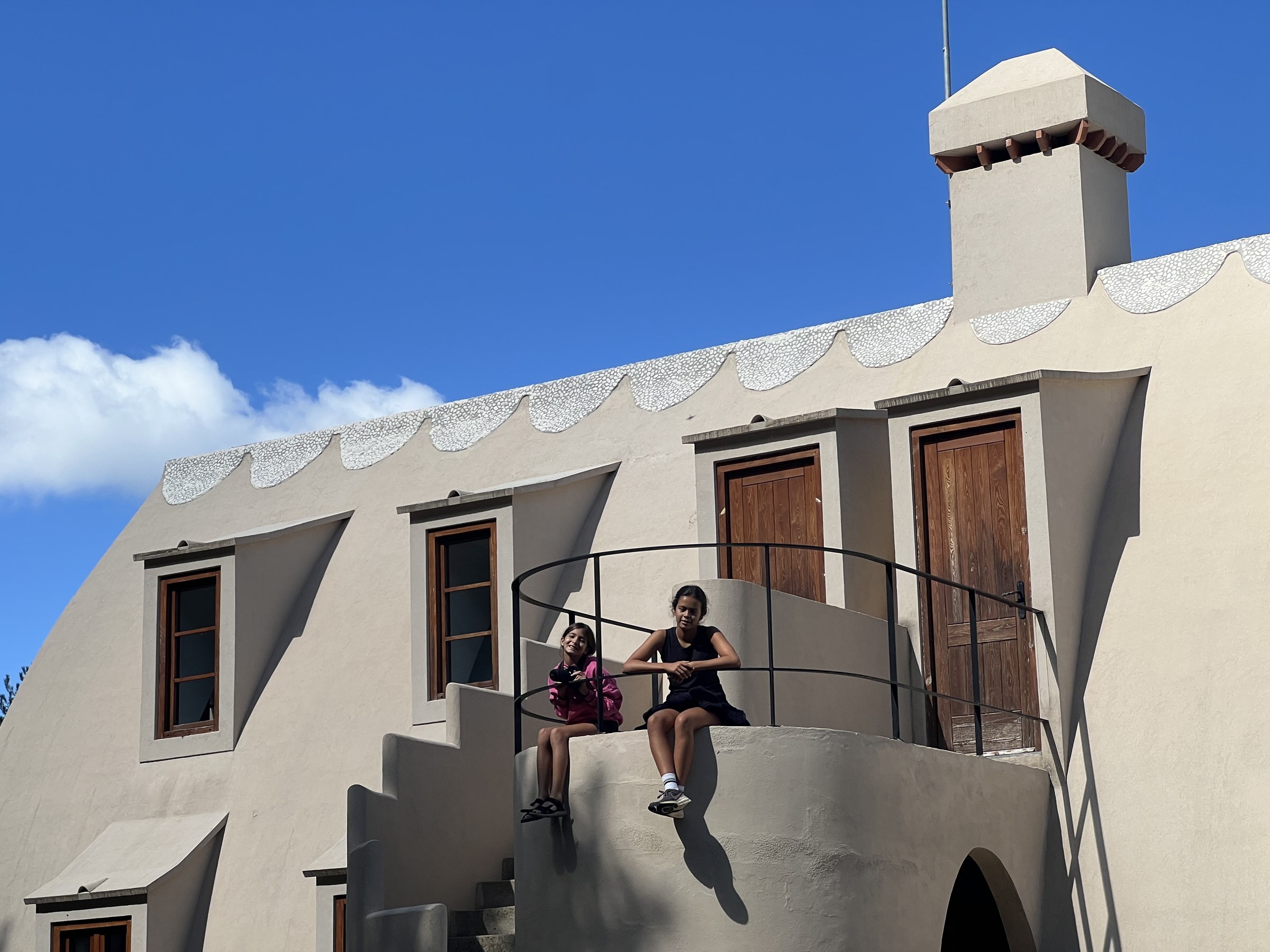 Two children sitting on the curved rendered landing of the Xalet del Catllaràs external stair beside an iron rail, with timber doors, dormer windows, a scalloped band of white pebbles along the roof ridge and a rendered chimney with a corbelled cap against a blue sky. RISE Design Studio.