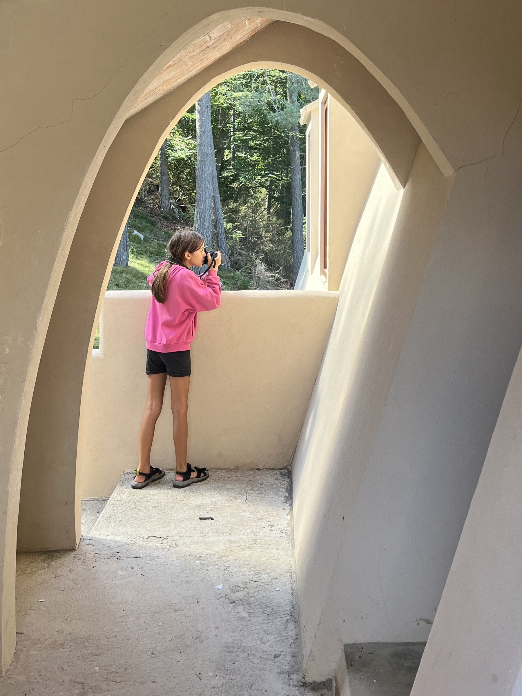 Inside the arched passage beneath the Xalet del Catllaràs external stair, a child in a pink top photographs the pine woods through a parabolic arch, with the sloping rendered roof-wall and a dormer window visible to the right. RISE Design Studio