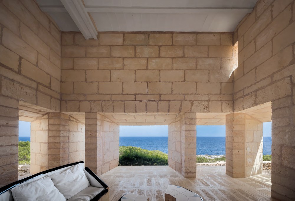 Deep splayed openings in the thick marés stone walls of the living room at Can Lis, Mallorca, framing the sea and coastal scrub with no visible window frames, and a tiled sofa in the foreground. RISE Design Studio.