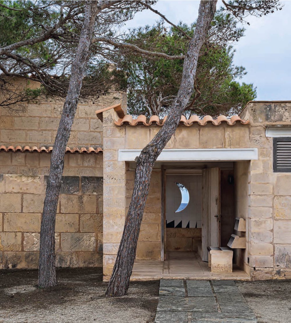 Entrance pavilion at Can Lis, Mallorca, with two pine trees in front of a marés sandstone wall and tiled roof, and a crescent-moon opening in white tiles visible through the open doorway. RISE Design Studio.