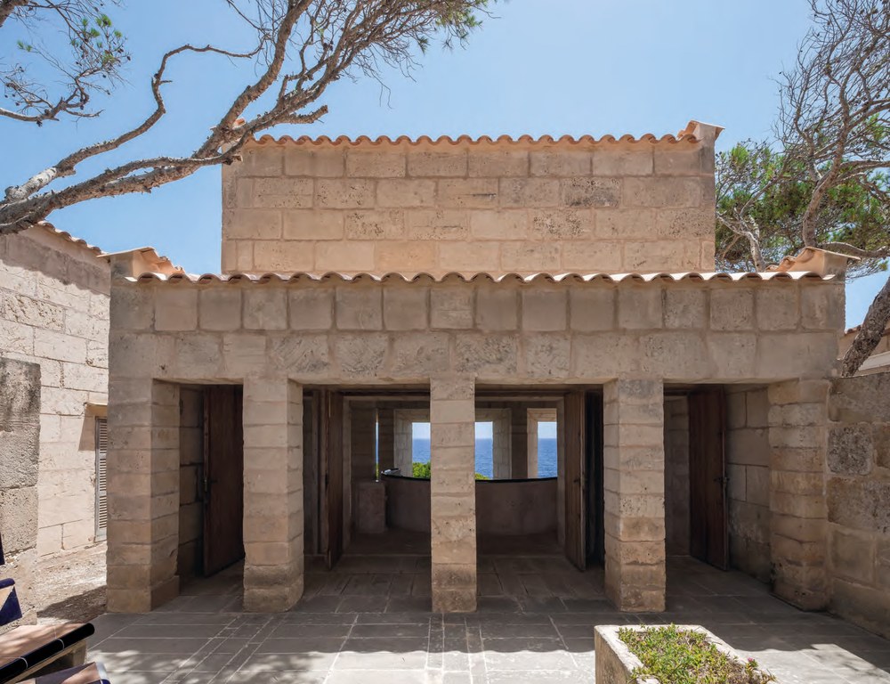 View from inside Can Lis through two wide openings to a paved courtyard, stone columns, terracotta tiled roofs and the deep blue Mediterranean beyond, on a bright sunny day in Mallorca. RISE Design Studio.