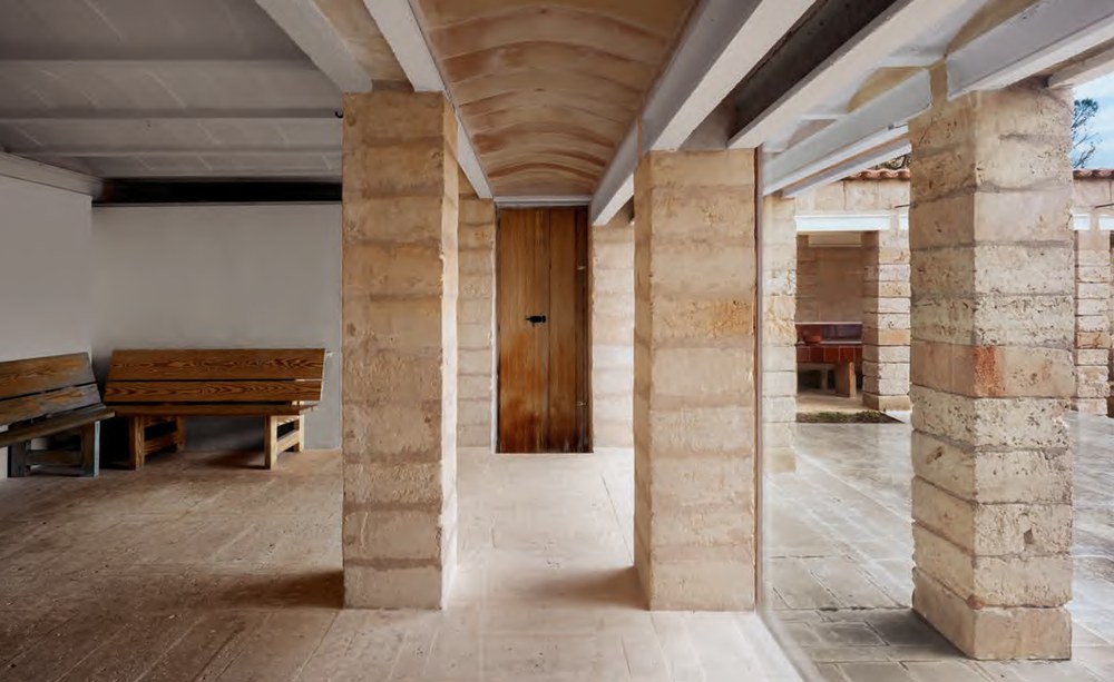 Interior at Can Lis by Jørn Utzon with stacked marés stone columns, a timber door, wooden benches and a curved ceramic ceiling between concrete beams, opening to a courtyard beyond. RISE Design Studio.