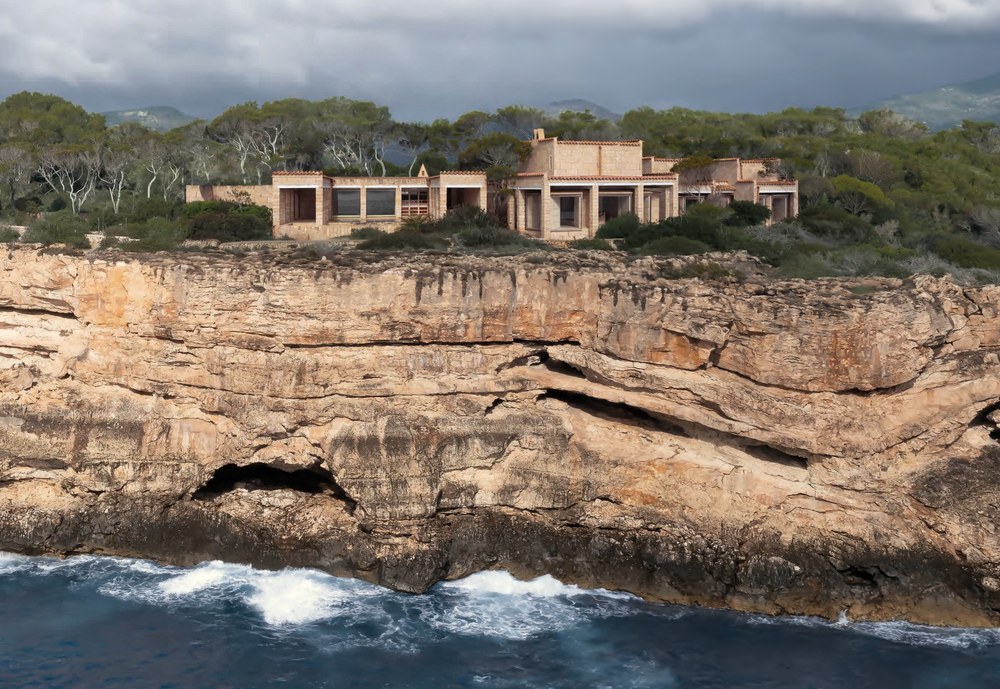 Can Lis by Jørn Utzon seen from the sea, a row of marés stone pavilions with tiled roofs along a cliff edge near Portopetro, Mallorca, with waves breaking below and pine trees and hills behind. RISE Design Studio.