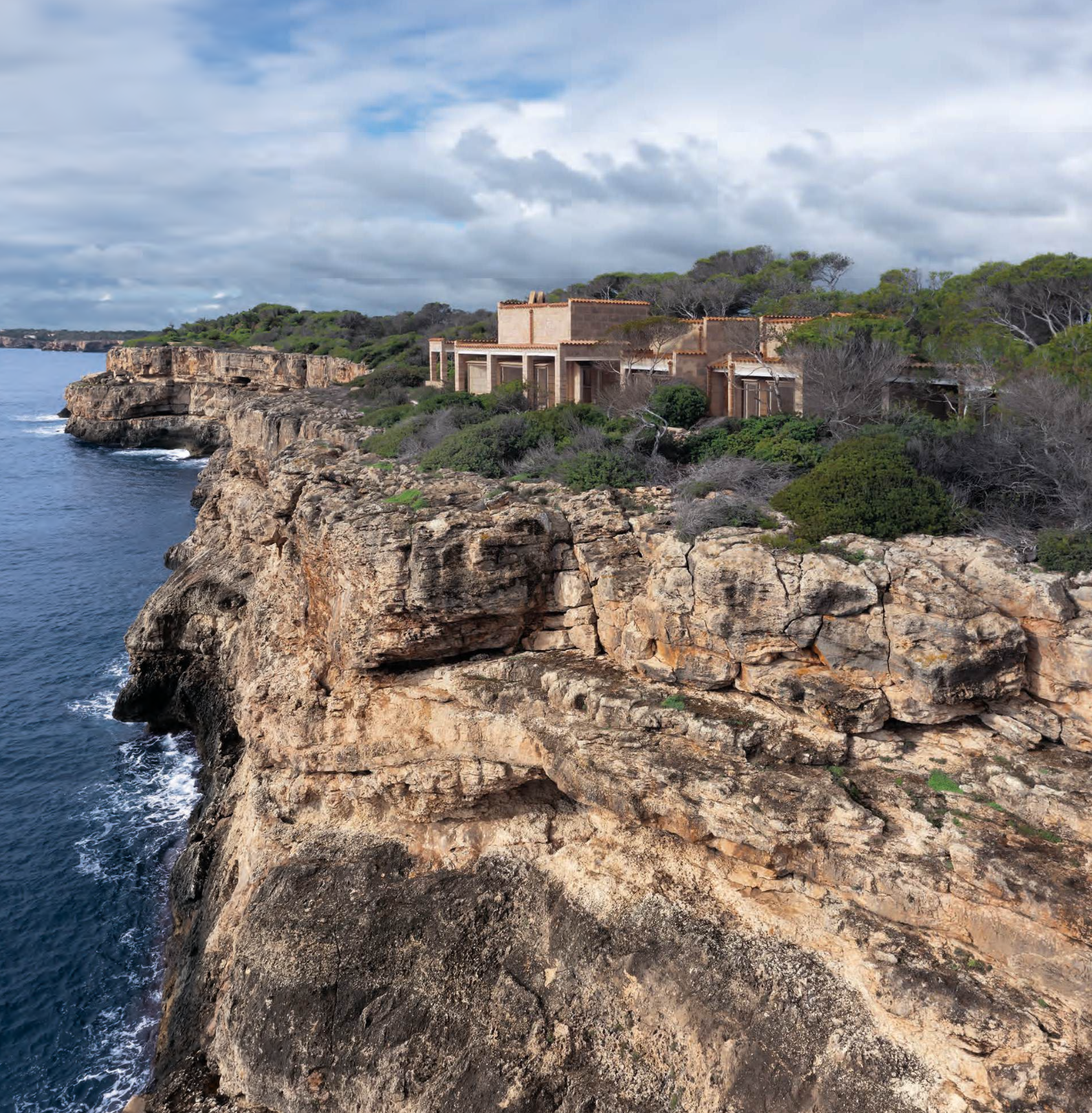 Aerial view of Can Lis by Jørn Utzon on a limestone cliff near Portopetro, Mallorca, with its marés stone pavilions and terracotta tiled roofs set among pines above a dark blue sea under cloudy skies. RISE Design Studio.
