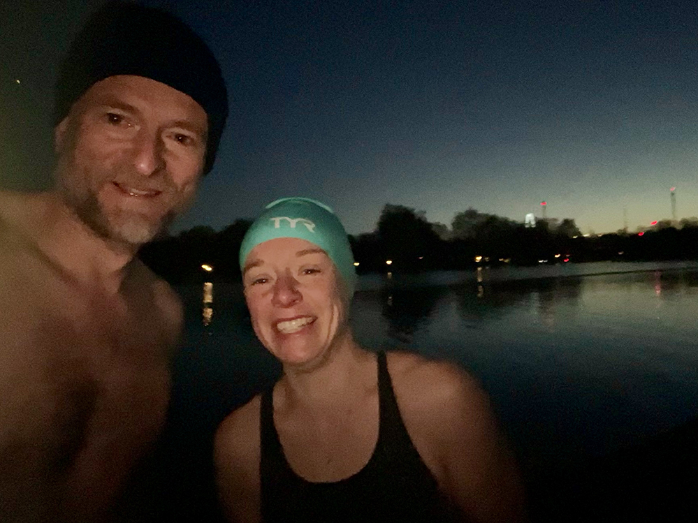 Two winter swimmers standing beside the Serpentine at dawn after a cold-water dip. The pair are smiling in hats and swim gear, with the lake and soft reflections of park lights behind them. The image captures the clarity, resilience and calm associated with early-morning cold-water swimming.