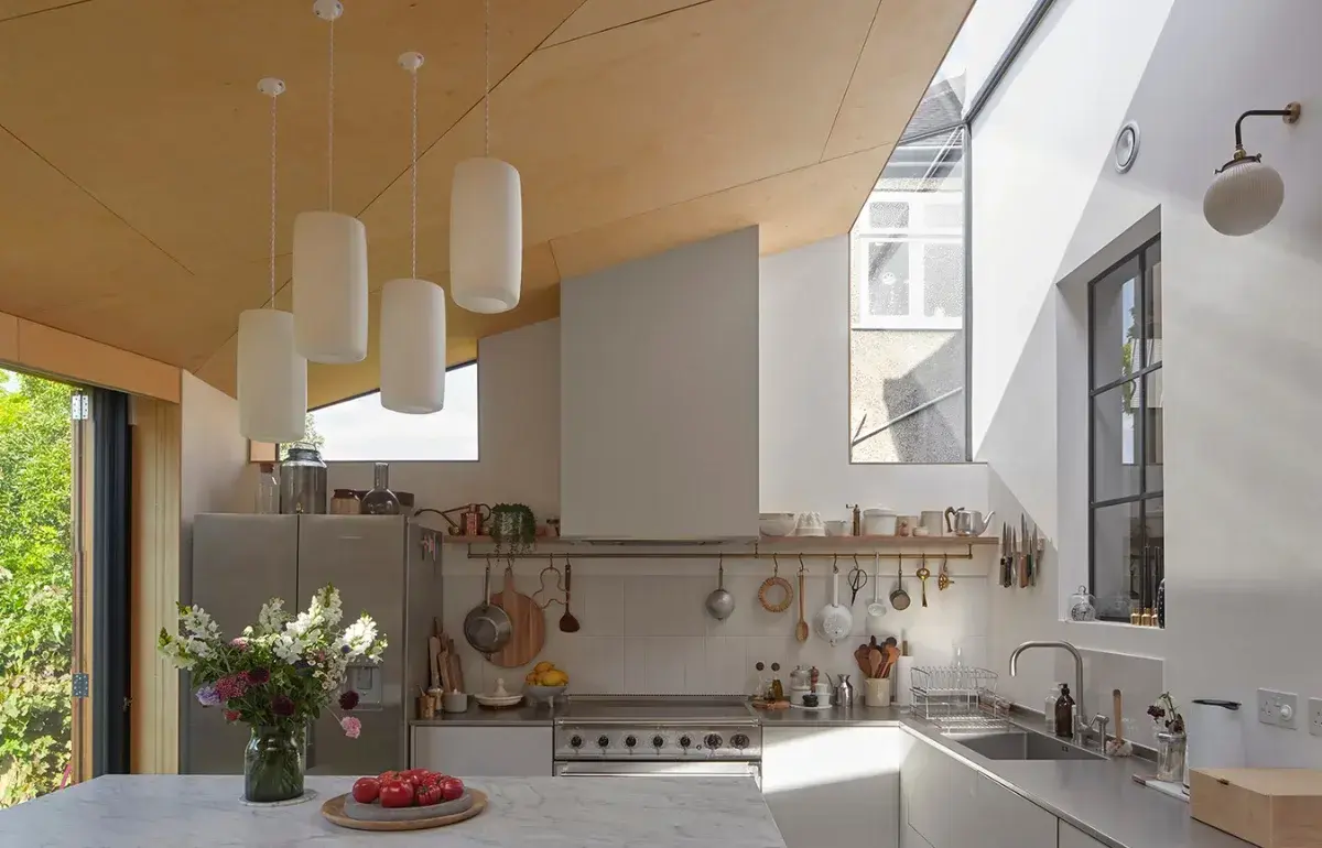 Douglas fir-lined kitchen at Douglas House in Kensal Rise by RISE Design Studio, with white pendant lights, a marble island, a range cooker and clerestory skylights drawing daylight across the timber ceiling toward sliding doors that open onto the garden.