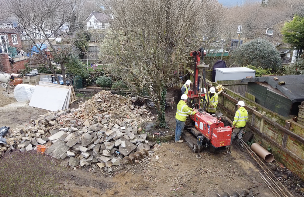 Builders operating piling rig on residential construction site during groundworks, with rubble and foundation materials visible around the area.