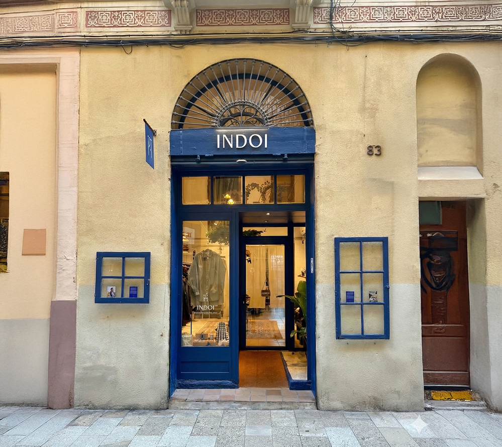 Exterior view of a slow fashion boutique, INDOI, in Barcelona designed by RISE Design Studio, showing a restored arched stone opening, deep blue timber storefront, glass door and warm interior lighting revealing handcrafted clothing and objects inside the small retail space.