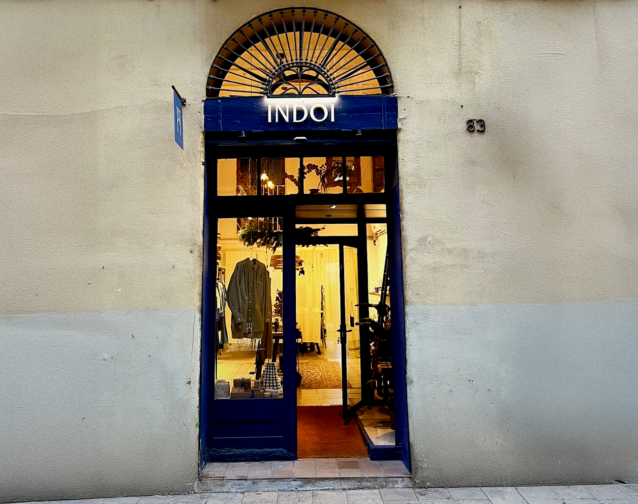 Exterior view of a slow fashion boutique, Indoi, in Barcelona designed by RISE Design Studio, showing a restored arched stone opening, deep blue timber storefront, glass door and warm interior lighting revealing handcrafted clothing and objects inside the small retail space.
